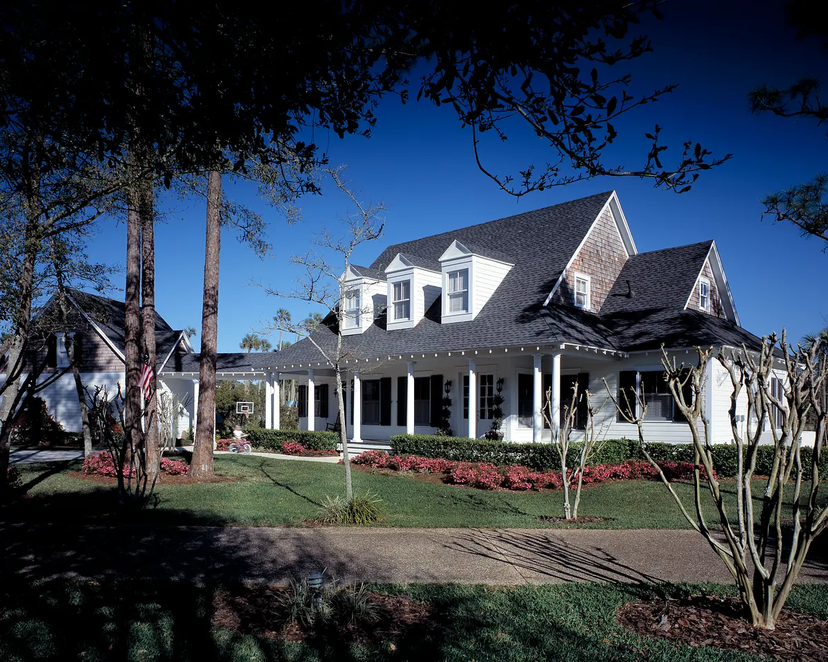 Front exterior of a two-story house with gabled roof, white siding, porch, flower beds, and trees