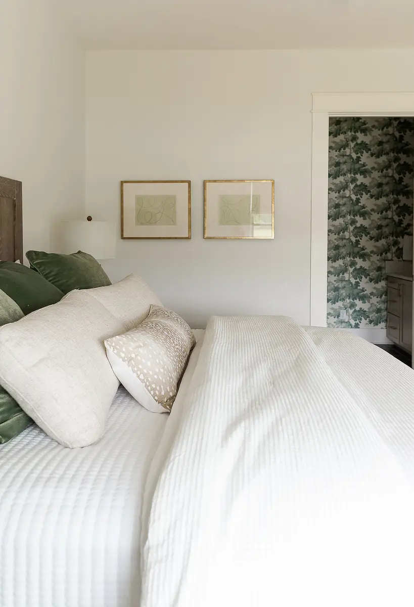Bedroom with large bed, white quilt, green cushions, framed art, and adjacent patterned wallpaper.