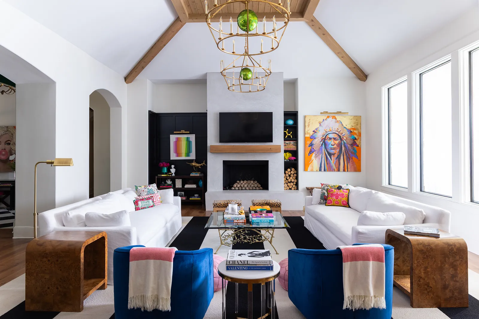 Living room with white sofas, blue chairs, coffee table, fireplace with TV, and large windows.