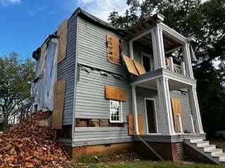 Exterior view of a two-story house with a collapsed wall, white columns, and a staircase.