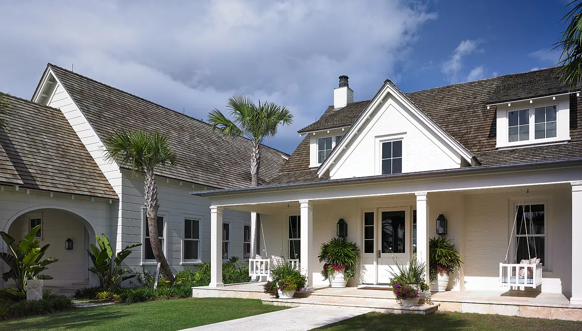 Exterior front view of a house with white brick, a gable roof, porch, and landscaped yard with palm trees.