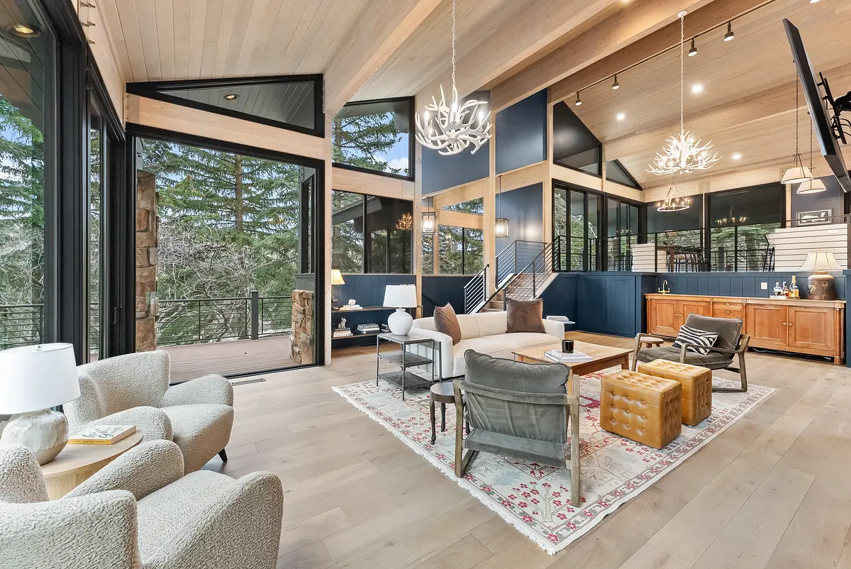 Living room with large windows, area rug, sofa, armchairs, ottomans, and wooden sideboard against blue paneling.