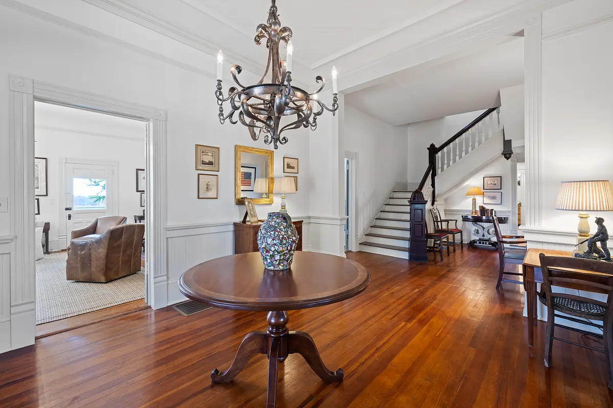 Foyer with round wooden table, colorful vase, staircase, and framed artworks on white walls.