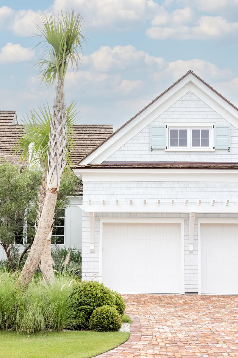 Garage with white doors, brick driveway, palm trees, and gabled roof with shutters.