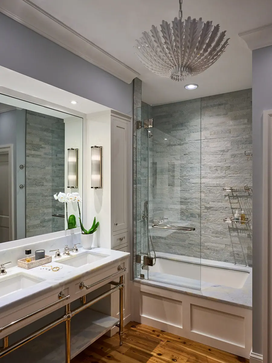Bathroom with double vanity, marble countertop, glass shower, tiled wall, and wall sconces.