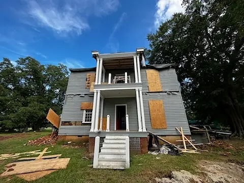 Exterior front of a two-story house with gray facade, boarded windows, white trim, and a staircase.
