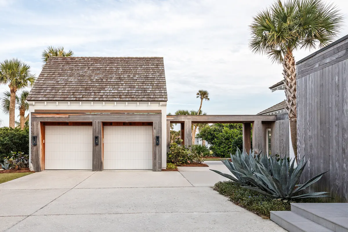 Garage with white doors, wood siding, shingled roof, palm trees, and concrete driveway.