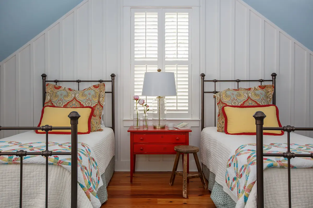 Bedroom with two metal frame beds, colorful quilts, red nightstand, table lamp, and wooden stool.