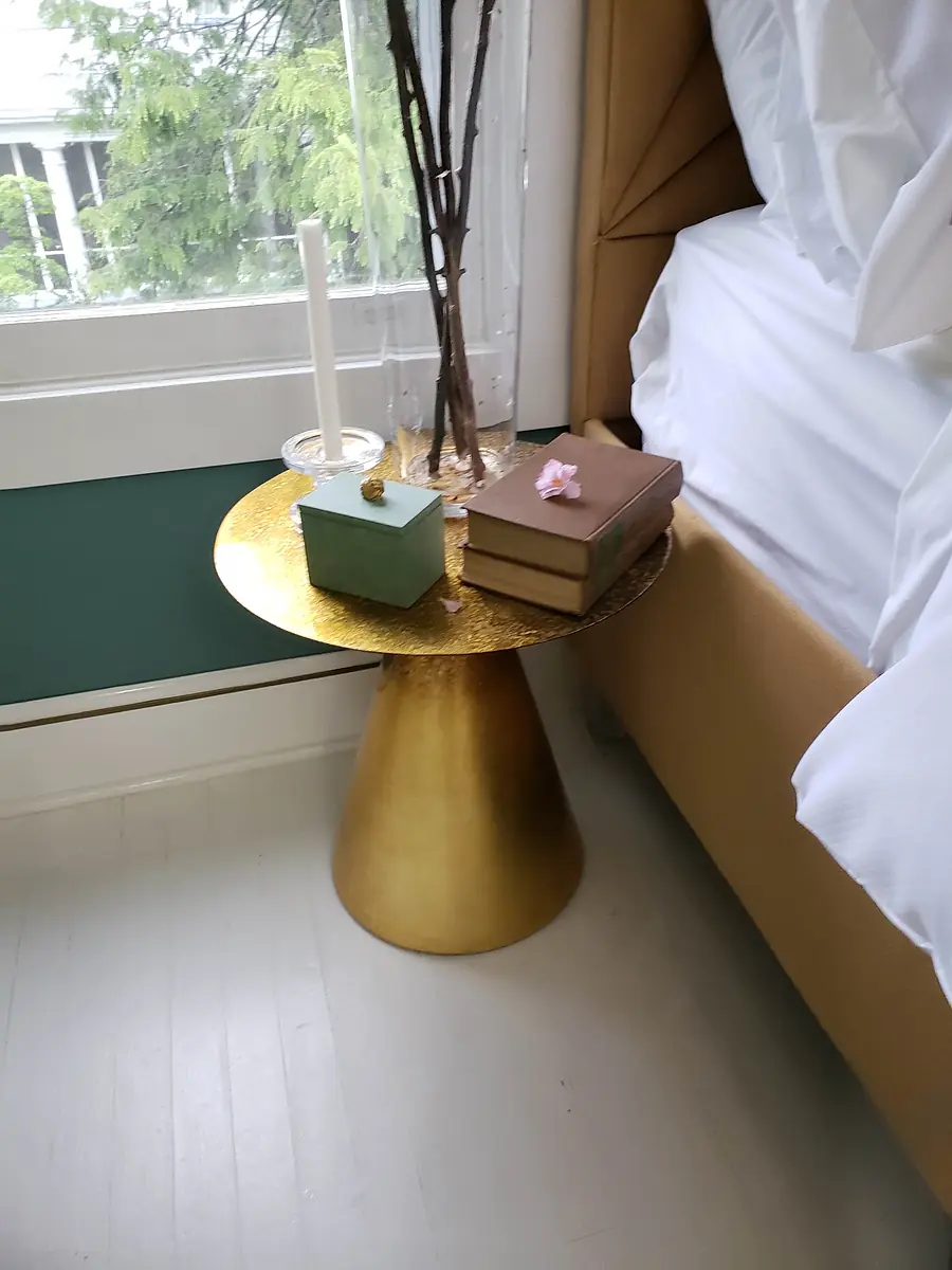 Bedroom with gold accent table, glass vase, candle, blue box, and stacked books near bed with white bedding.