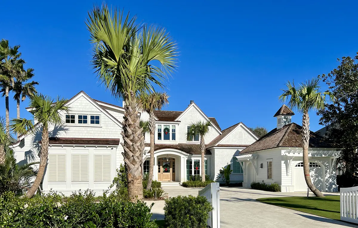 Front exterior of a house with white siding, palm trees, double doors, and a garage structure.