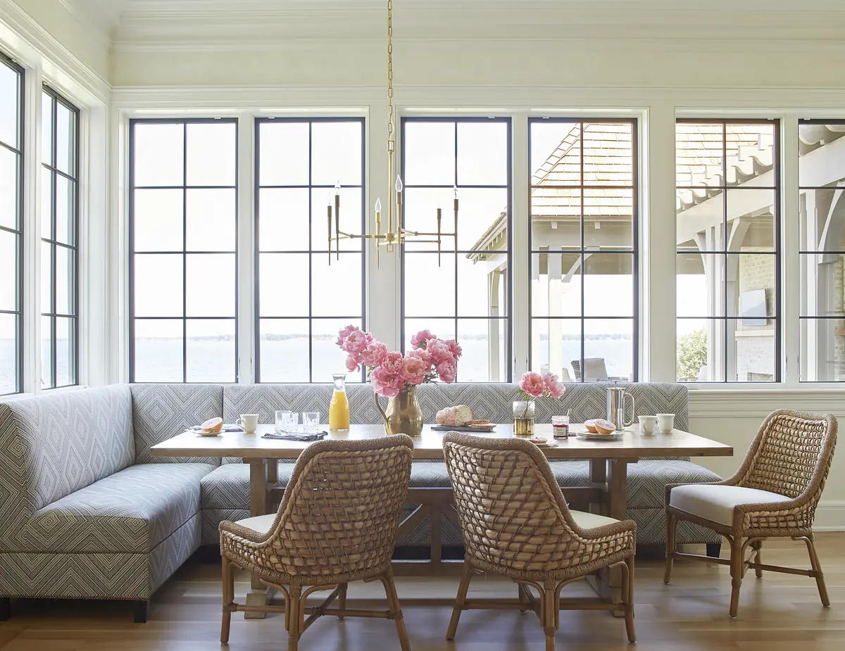 Dining room with wooden table, rattan chairs, L-shaped banquette, large windows, and floral centerpieces.