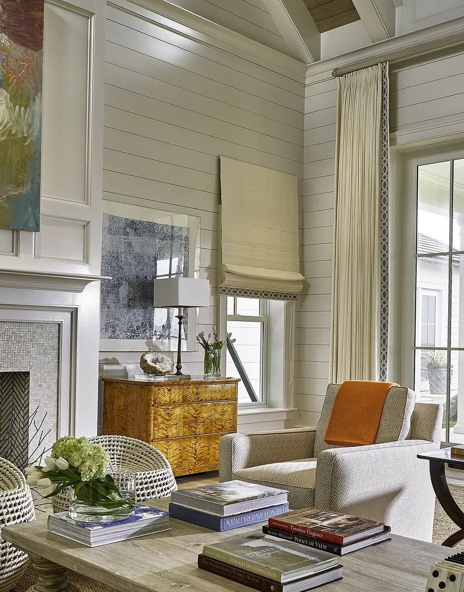 Living room with beige armchair, wooden side table, decorative books, large window, and fireplace with mosaic front.