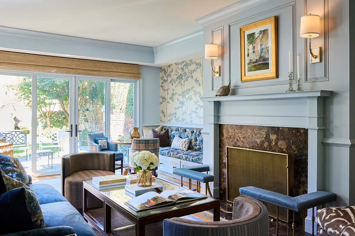 Living room with blue sofa, armchairs, coffee table, marble fireplace, floral wallpaper, and glass doors to outdoors.