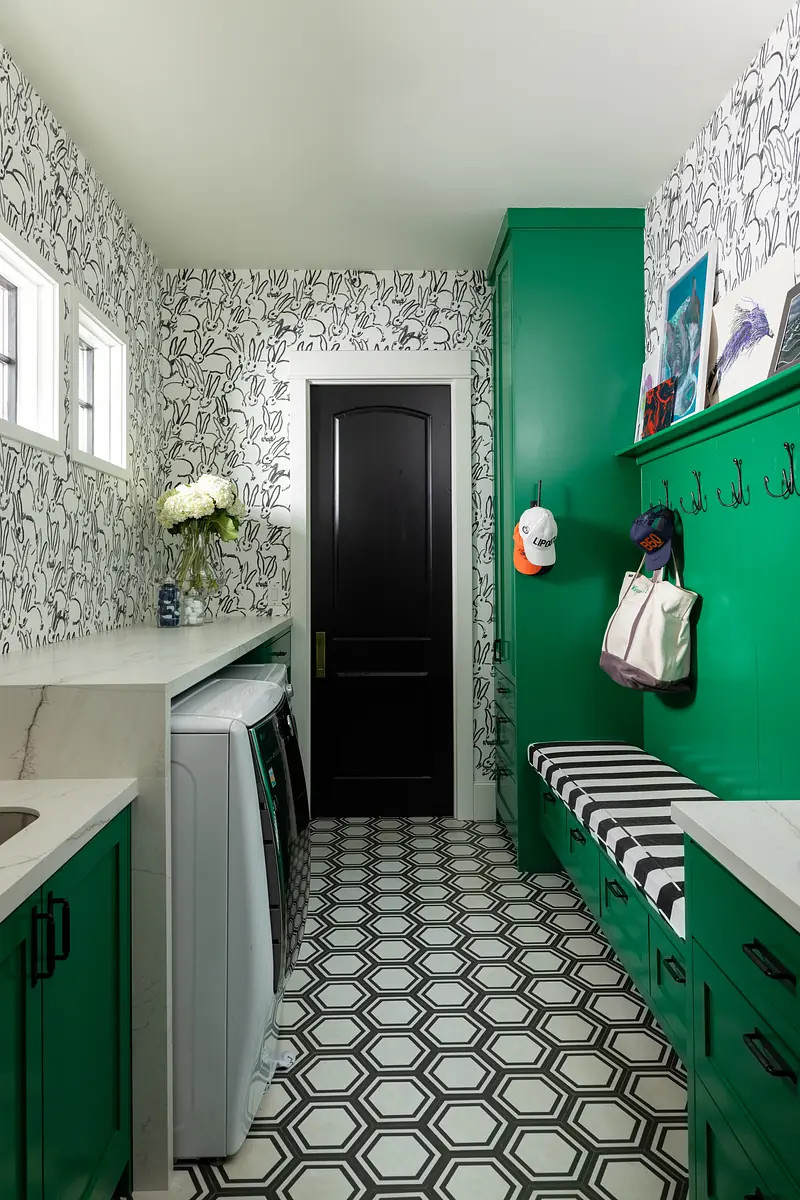 Laundry room with green cabinets, white countertop, washing machine, patterned floor, wall hooks, and bench.