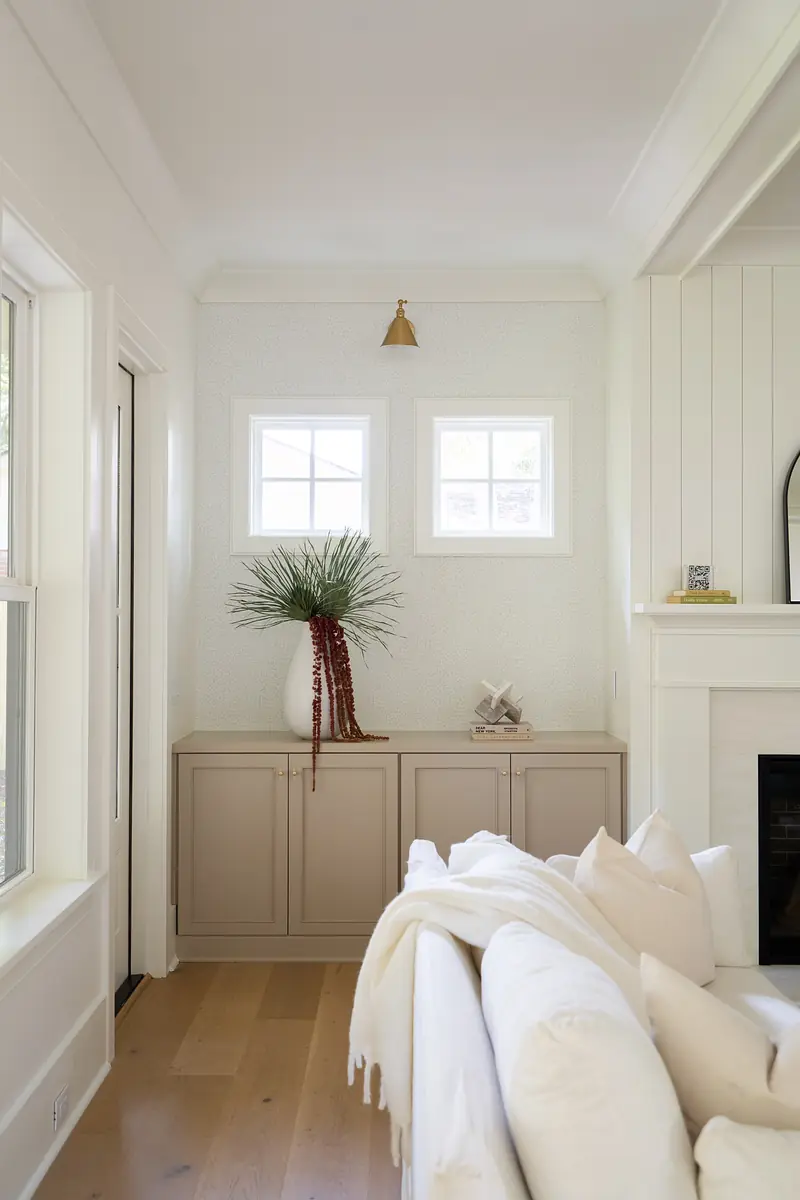 Living room with white sofa, beige cabinet, decorative vase, two windows, and a wall-mounted light fixture.