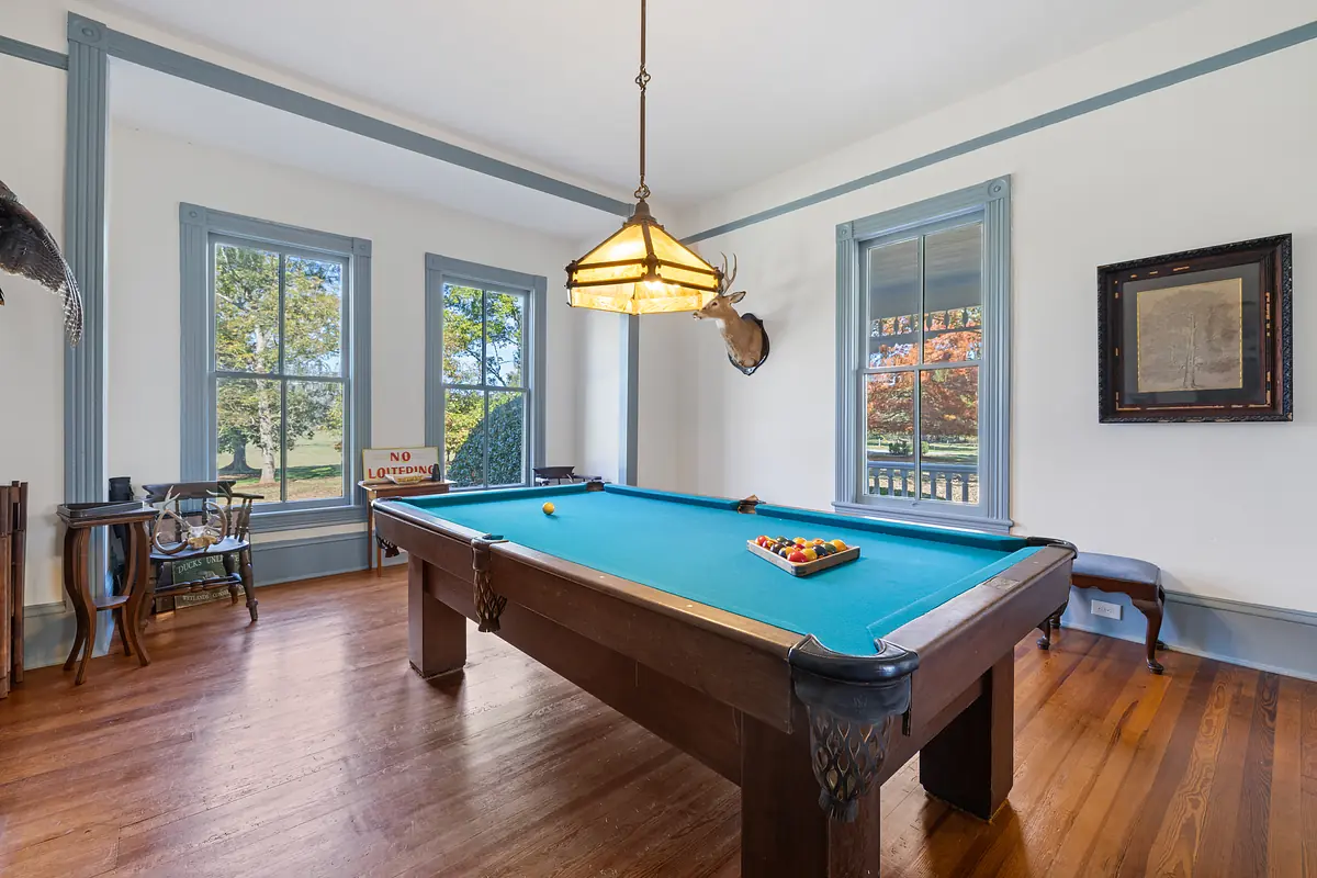 Room with billiard table, pendant light, large windows, framed artwork, and hardwood floor.