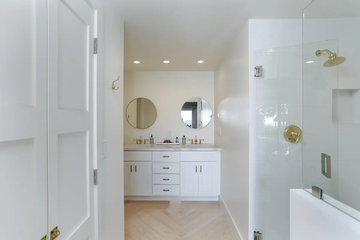 Bathroom with white vanity, gold fixtures, two mirrors, and glass shower enclosure on light wood floor.