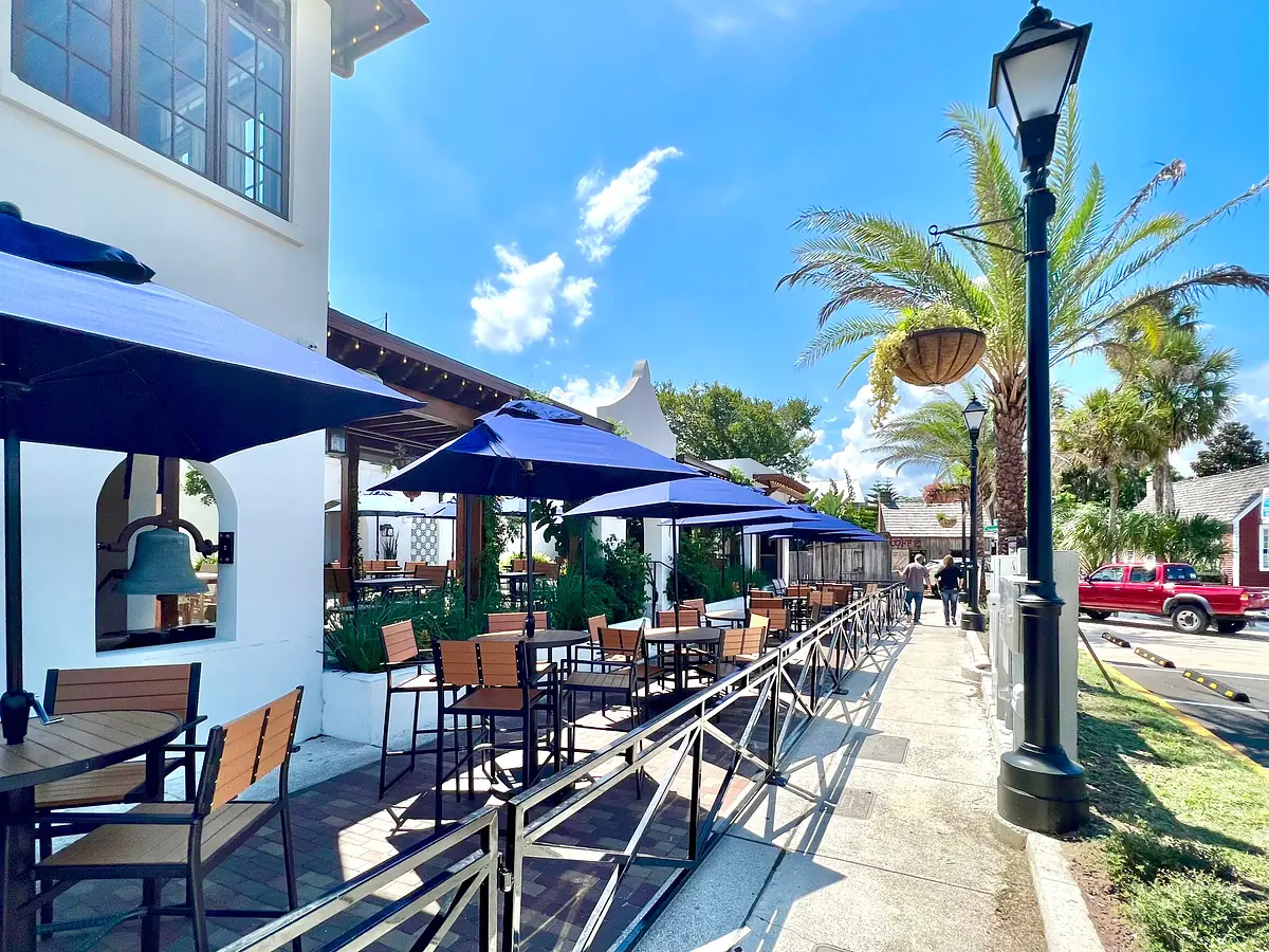 Outdoor patio with wooden tables, chairs, blue umbrellas, and landscaped foliage.