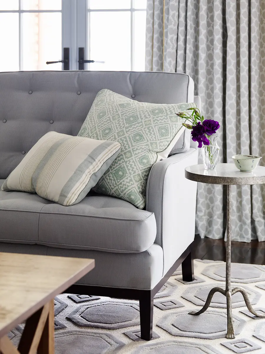 Living room with gray sofa, round side table, patterned rug, and window with drapes providing natural light.