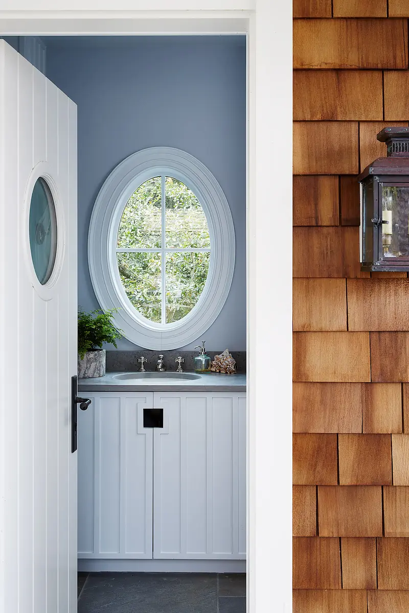 Powder room with circular window, white cabinet, sink, and light blue walls, featuring partially visible door and wood siding.