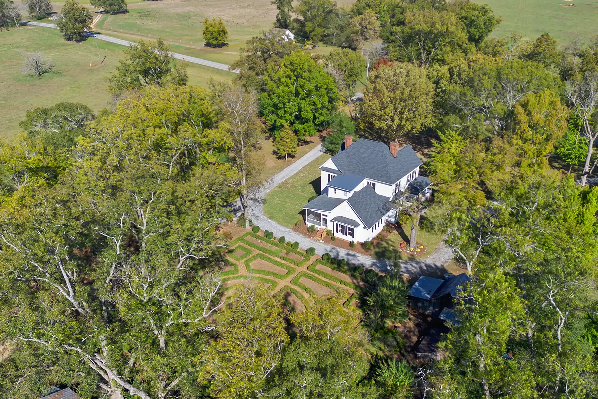 Aerial view of a two-story house with gray roof, white siding, porch, and geometric hedge garden.