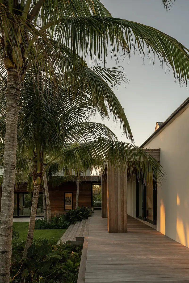 Exterior front with wooden walkway, palm trees, greenery, white wall, and wooden pergola structure