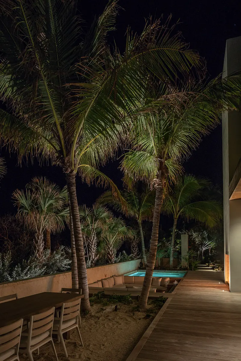 Outdoor patio with wooden dining table, chairs, palm trees, and swimming pool visible at night