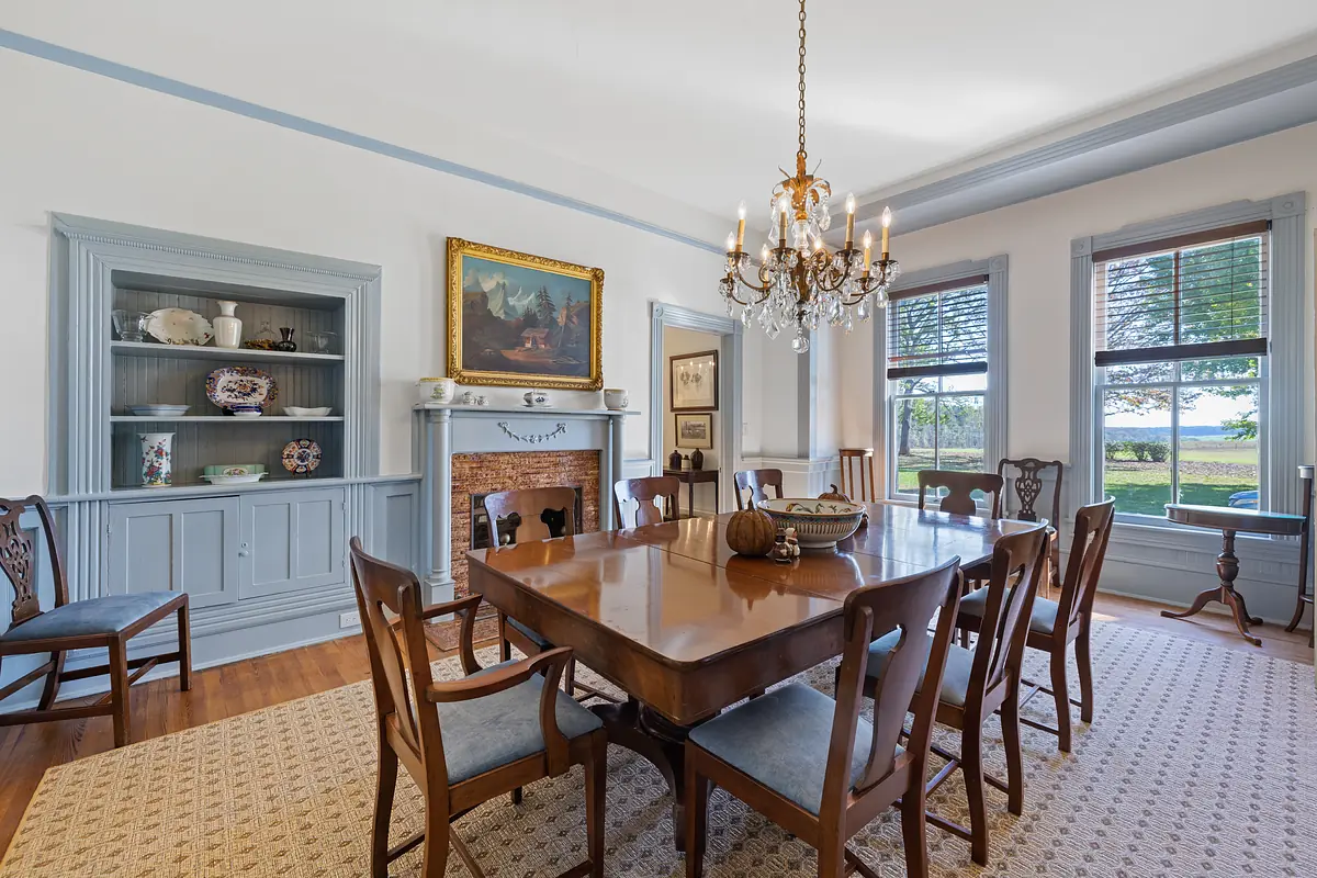 Dining room with large wooden table, eight chairs, chandelier, china cabinet, and large windows.