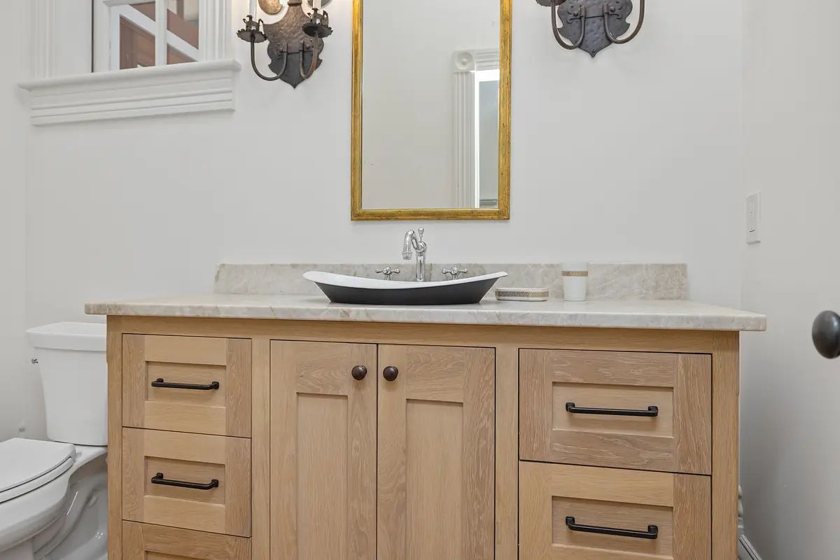 Powder room with wood vanity, beige countertop, black vessel sink, round gold-framed mirror, and two sconces.