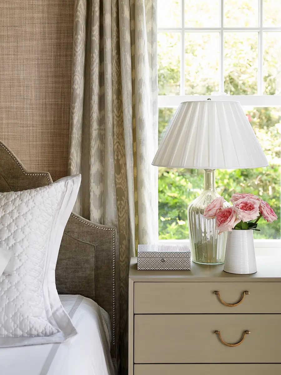 Bedroom corner with upholstered headboard, white bedding, nightstand, glass lamp, and pink roses.