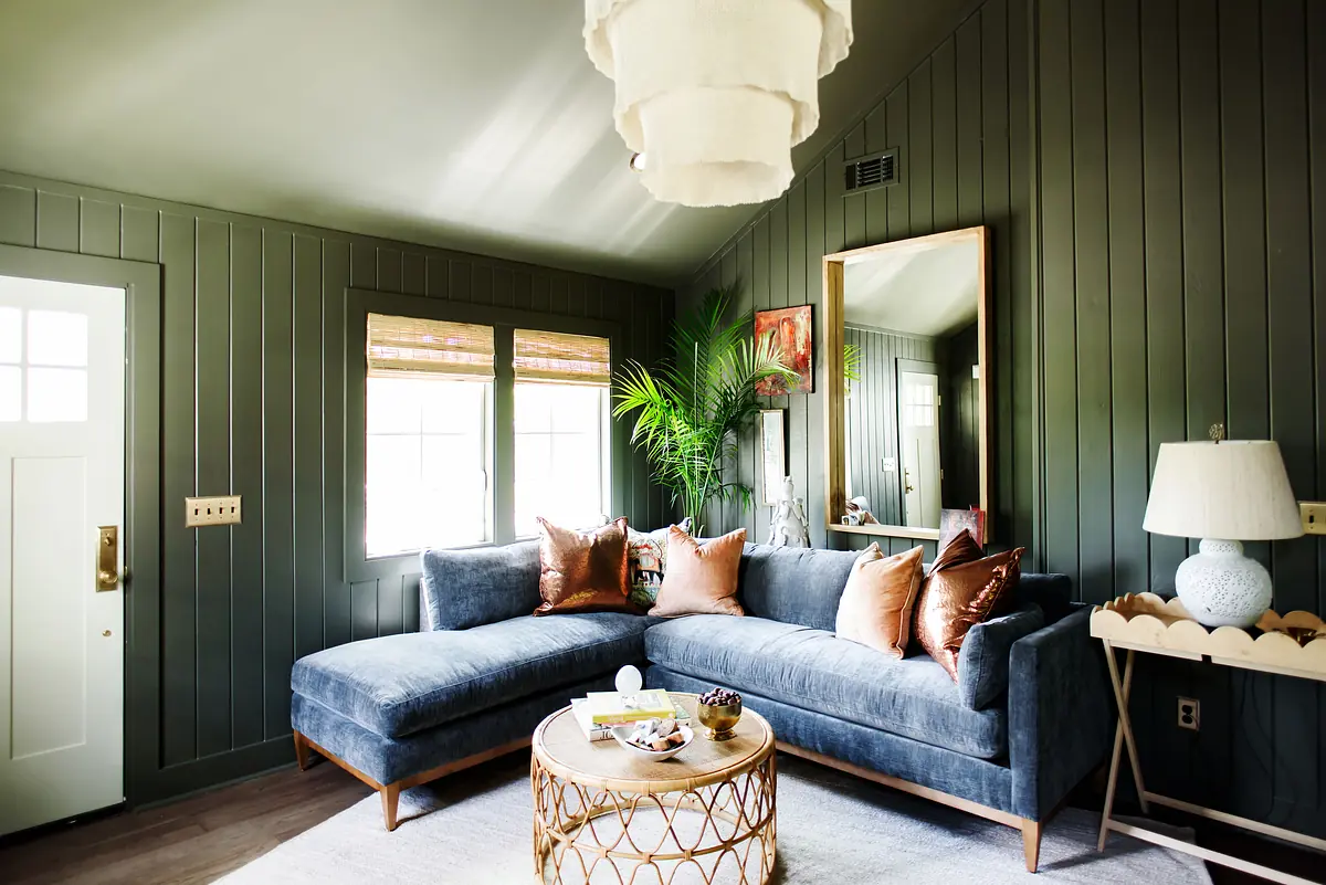 Living room with blue sectional sofa, round coffee table, floor lamp, and greenery against dark green paneled walls.