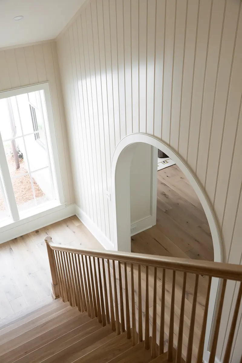 Hallway with arched doorway, vertical paneling, wooden staircase, and large windows for natural light.