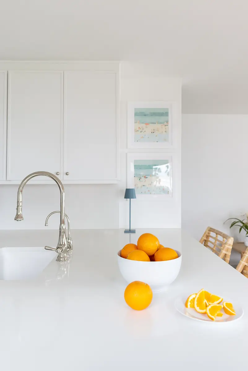 Kitchen with white countertop, stainless steel faucet, artwork, bowl of oranges, and wooden chairs.
