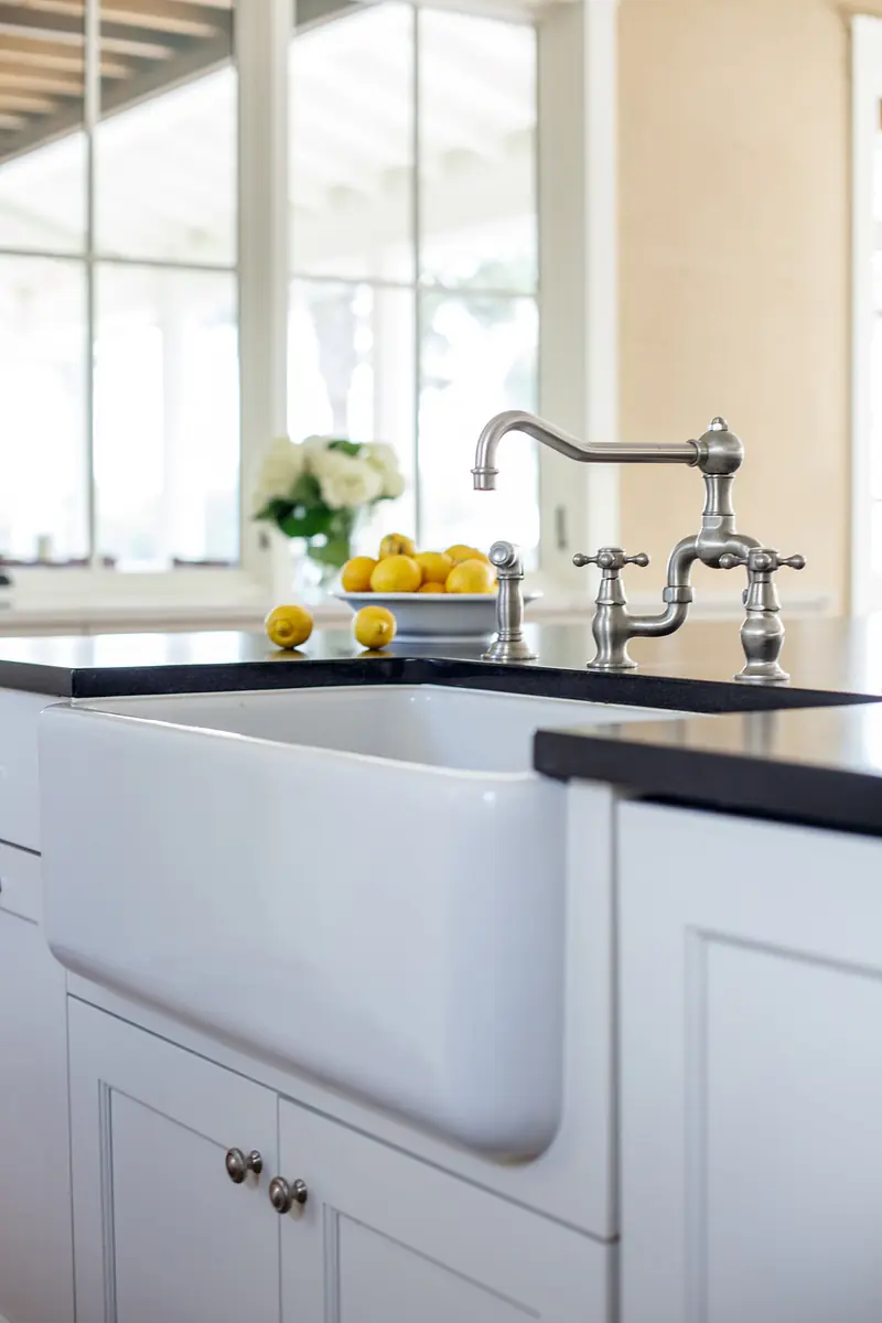 Kitchen with farmhouse sink, white cabinetry, black countertop, bowl of lemons, and flowers with natural light.