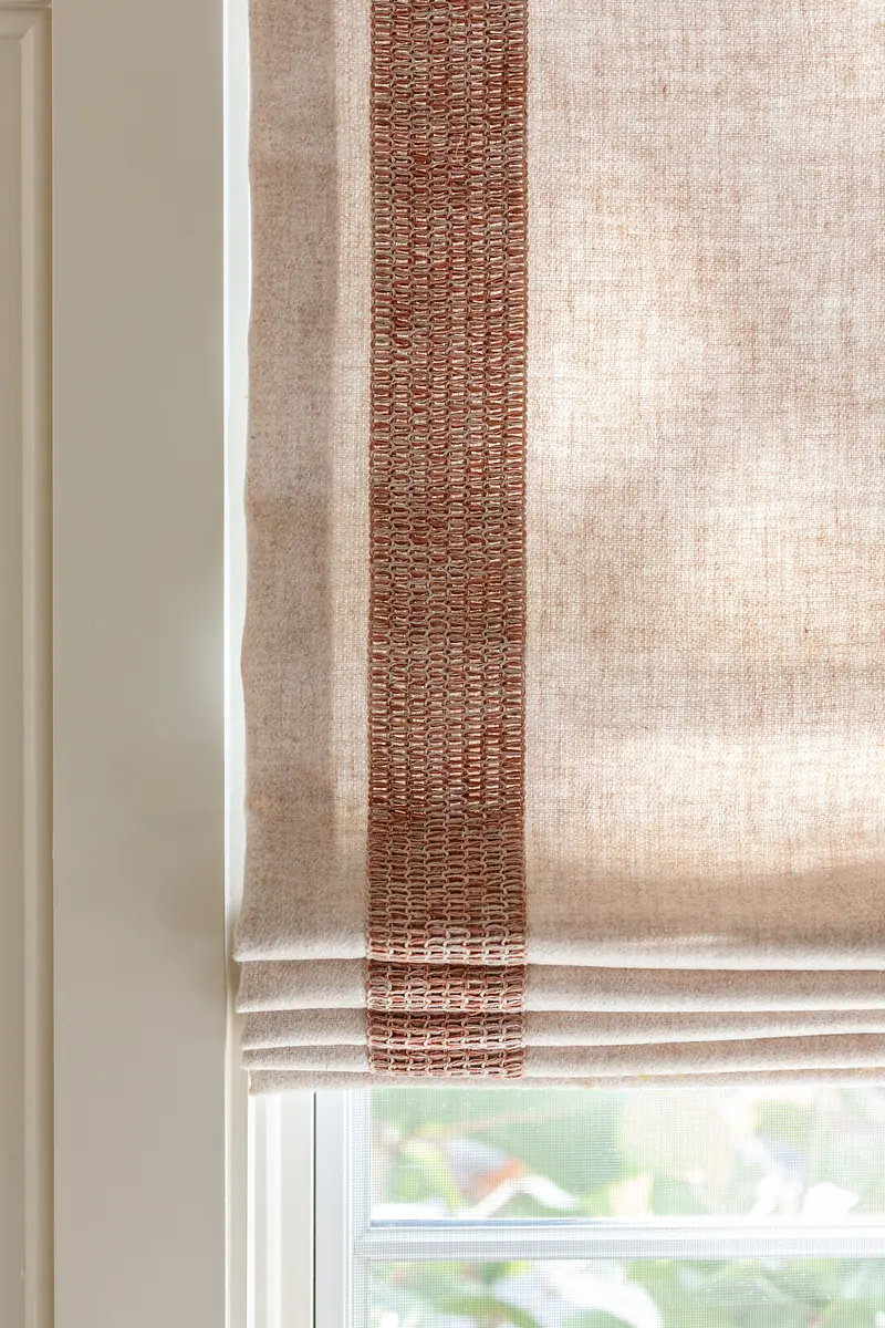 Fabric window treatment with beige material and a reddish woven stripe.