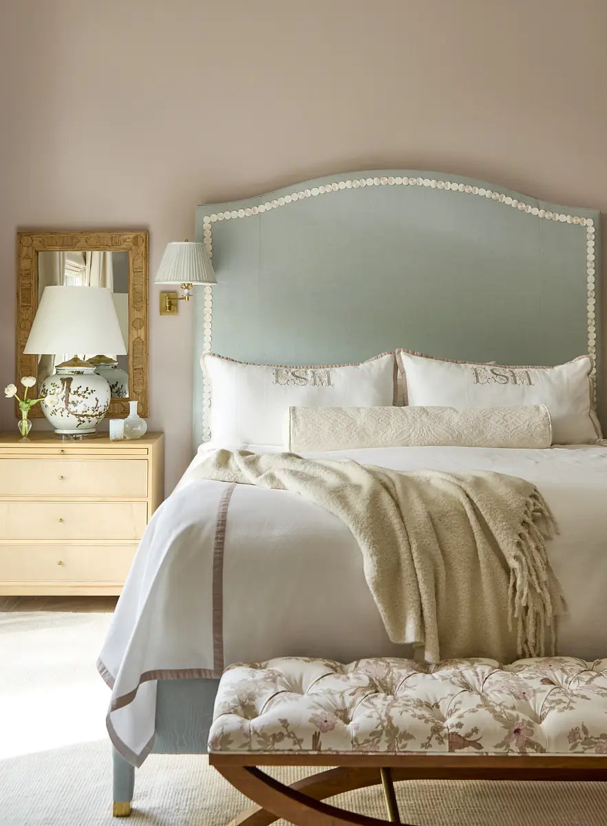 Bedroom with light blue headboard, white bedding, nightstand, lamp, mirror, and floral-patterned bench.