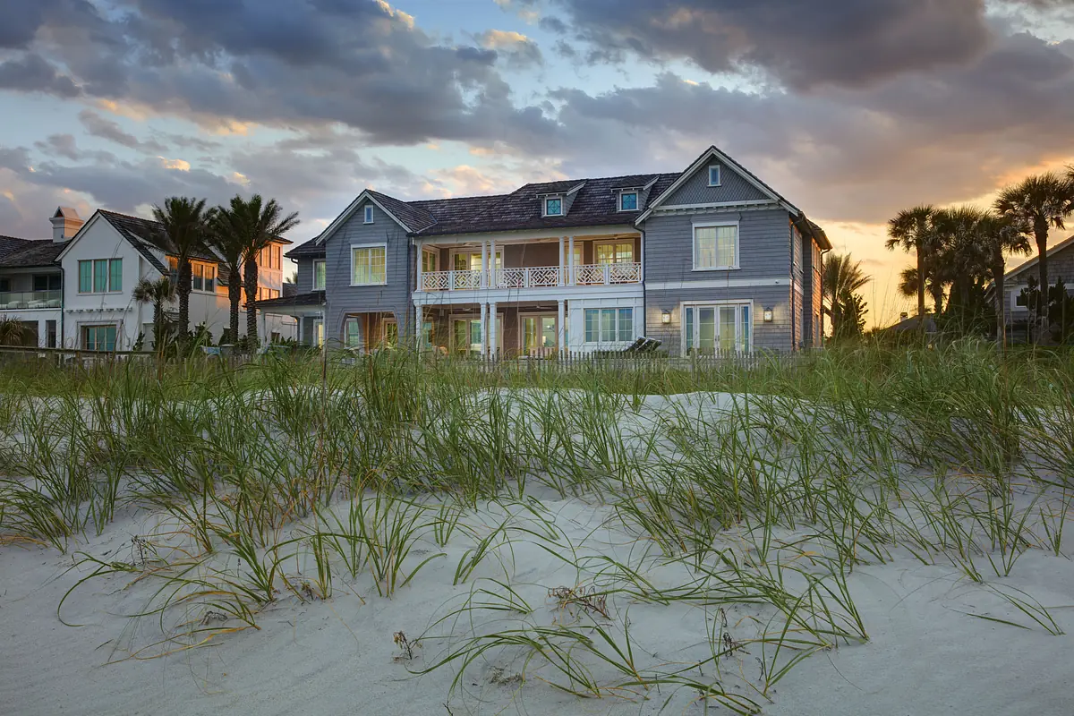 Coastal house with gray siding, large windows, balconies, and sandy beach with grass and palm trees.