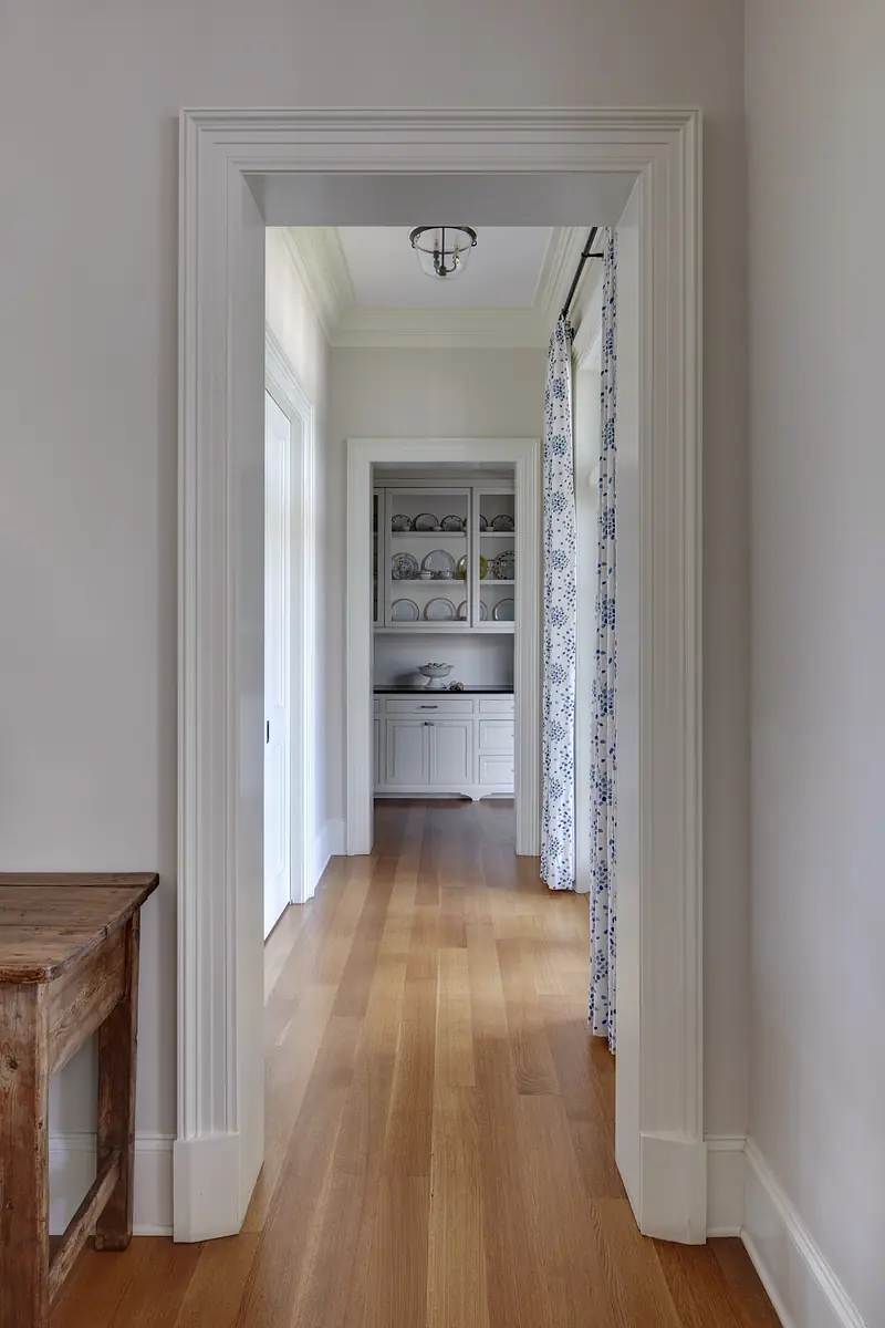 Hallway with light walls, hardwood flooring, wooden console table, and curtains leading to a pantry area with dishes.
