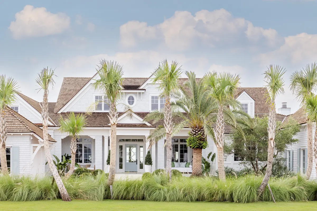Front exterior of a house with white siding, peaked roof, palm trees, and double doors with a porch.