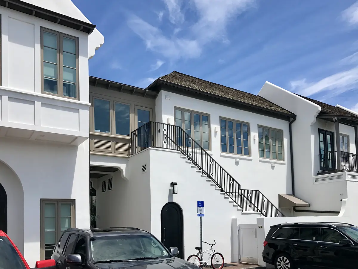 Exterior front of a building with white walls, large windows, stairs, and parked cars.