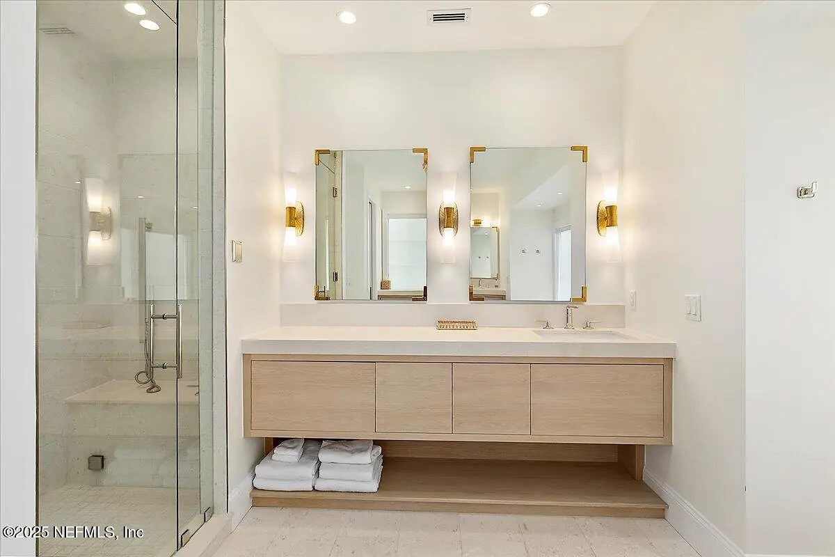 Bathroom with double sink vanity, mirrors, wall sconces, glass shower, and stacked towels.