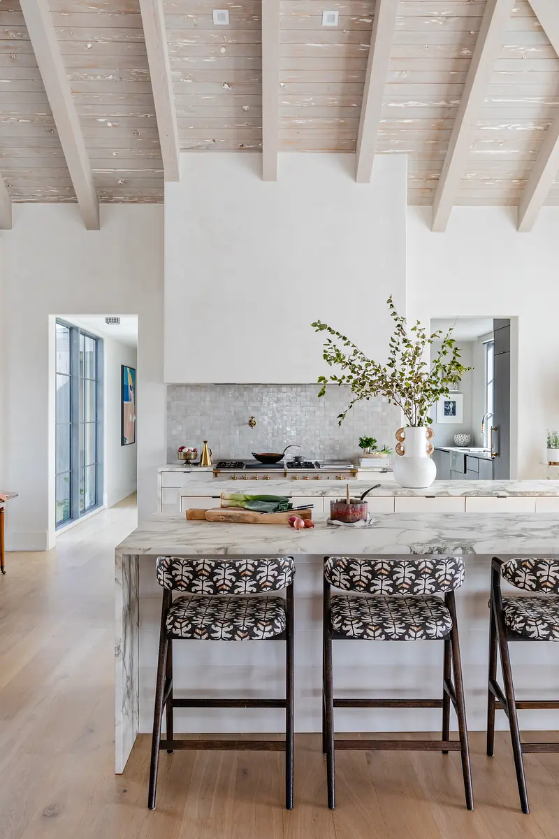 Kitchen with marble island, patterned bar stools, gray tile backsplash, and wooden beams with greenery arrangement.
