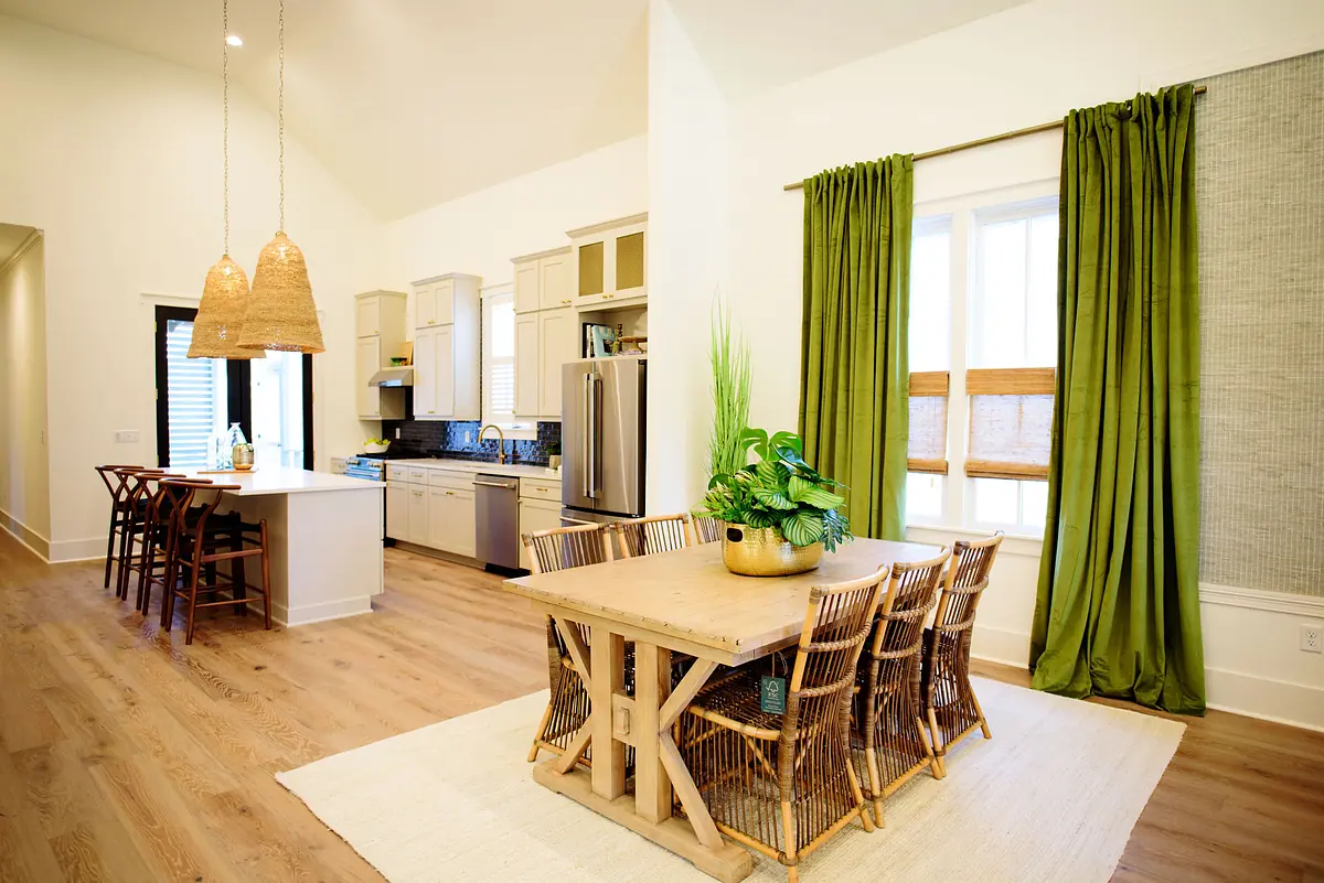 Dining room with wooden table, wicker chairs, greenery, pendant lights, large windows, and an open kitchen area.