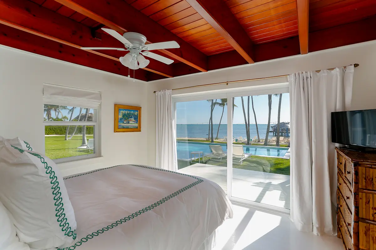 Bedroom with king-size bed, wooden dresser, TV, and views of pool and palm trees from windows