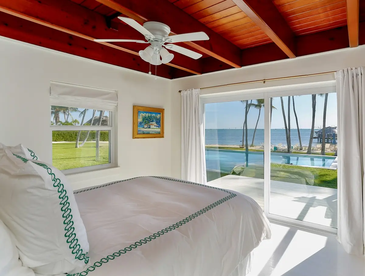 Bedroom with white bedding, ceiling fan, large window, sliding glass door, and wooden ceiling beams.