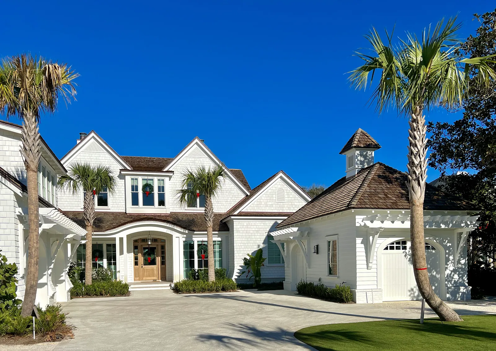 Front exterior of a house with light facade, gabled roofs, wooden door, palm trees, and a separate garage structure.