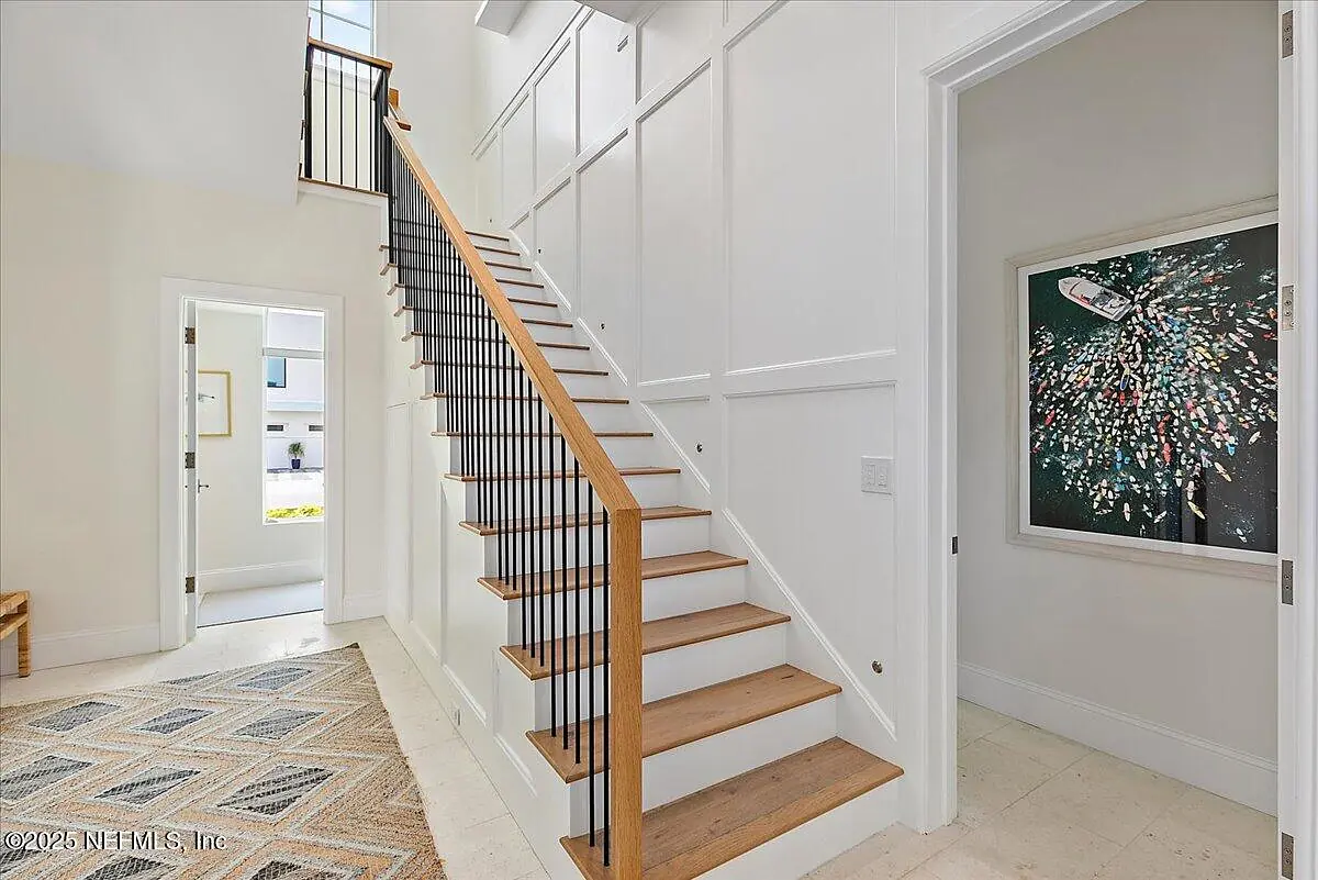 Foyer with staircase, white wall paneling, area rug, doorway, and artwork on the wall.