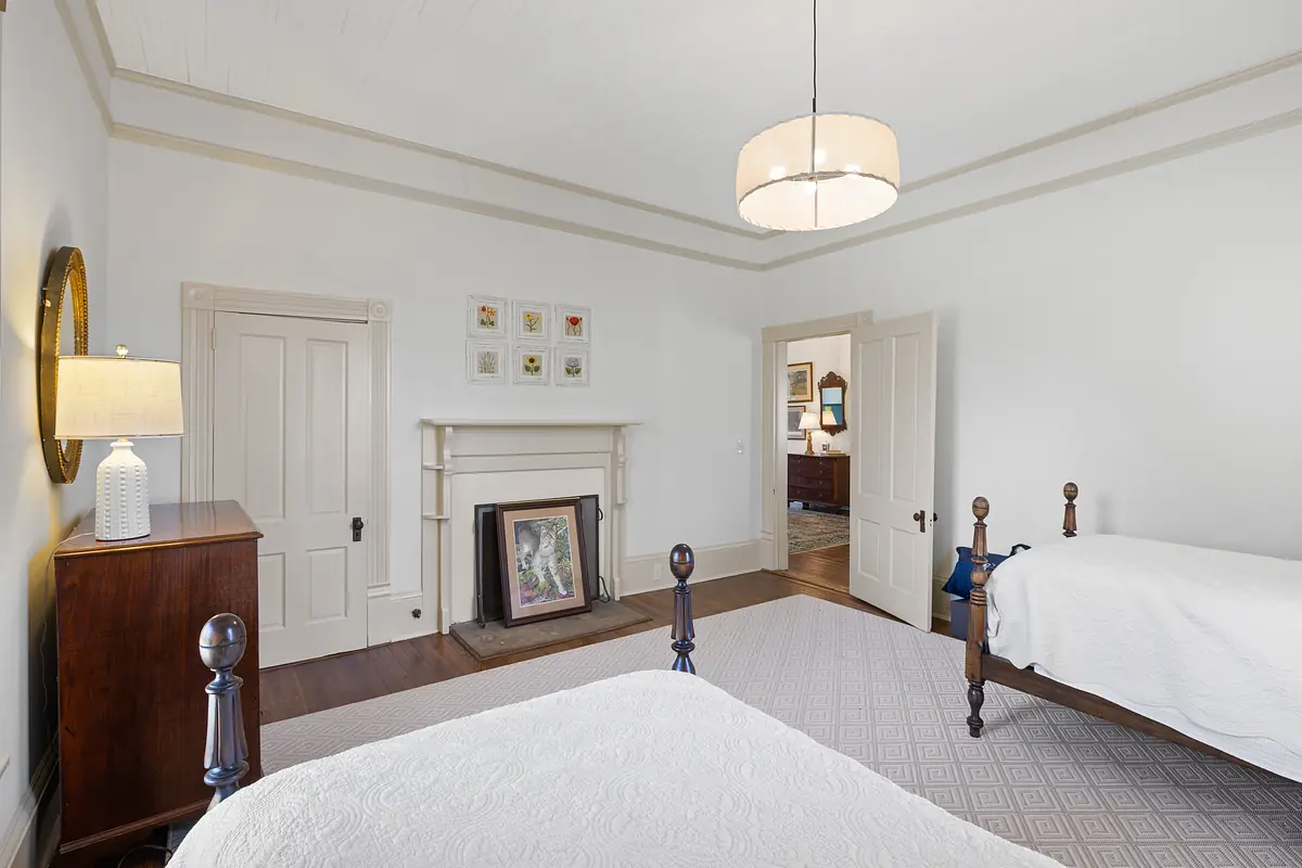 Bedroom with two wooden beds, white quilts, nightstand, lamp, fireplace, and beige walls.