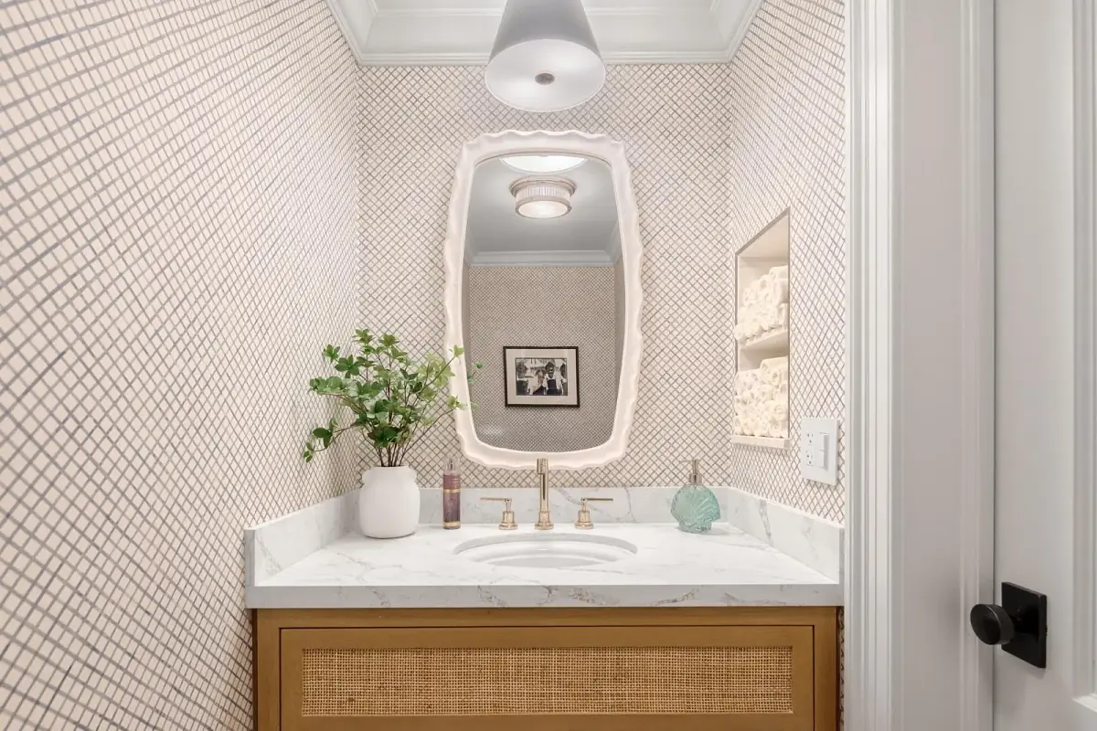 Powder room with marble vanity, wooden cabinet, round mirror, small plant, and patterned wallpaper.