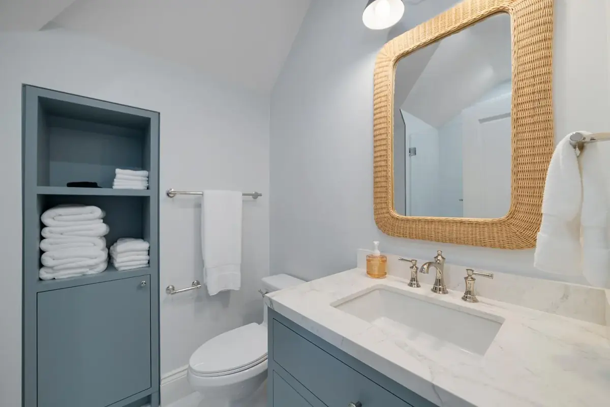 Bathroom with light blue walls, white marble countertop, woven mirror, towels, and toilet.